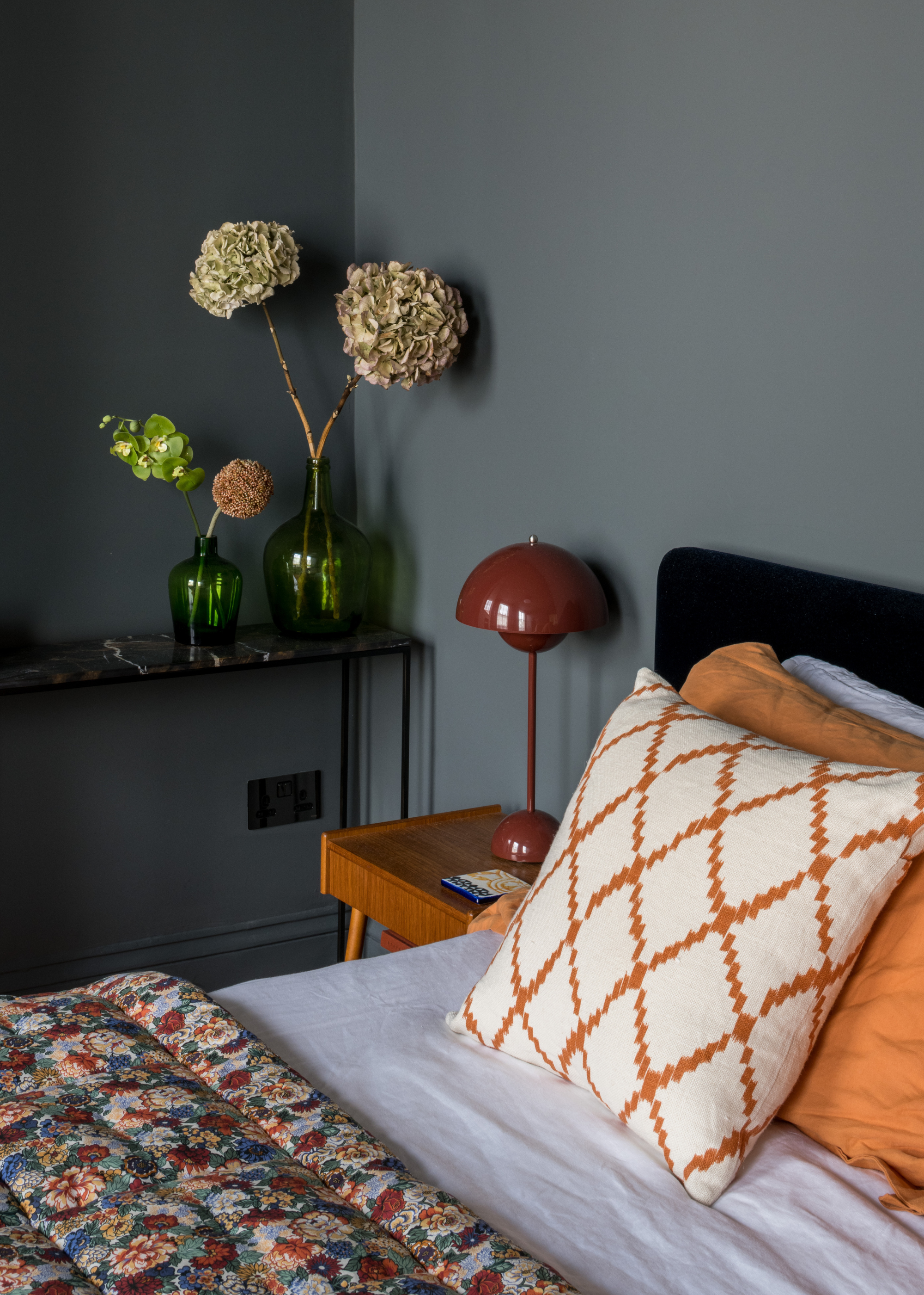 A dusty blue bedroom with a bed featuring colorful pillows, a patterned quilt, a wood bedside table with a dome lamp and a side table with vases of hydrangeas