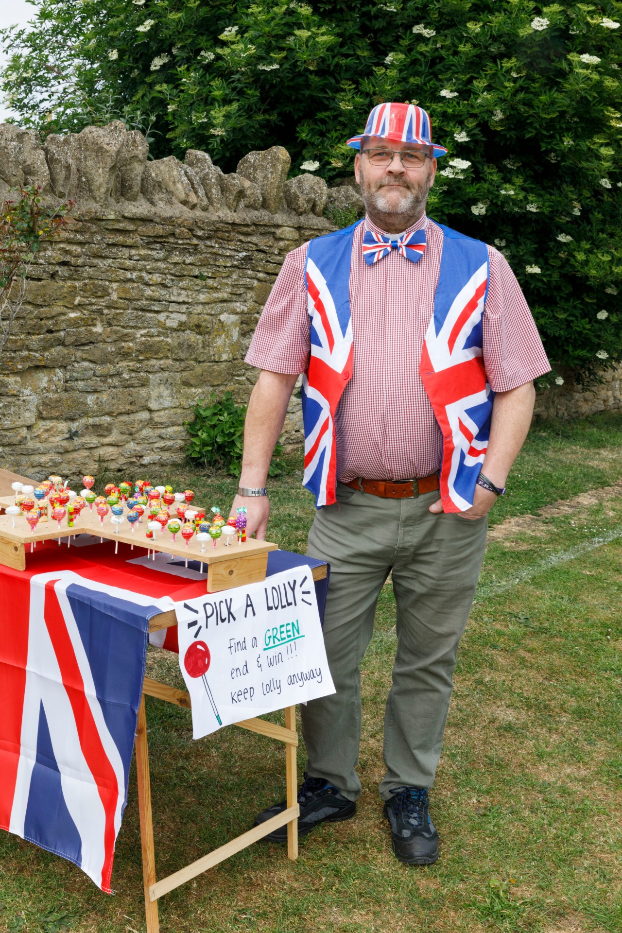 Man beside table draped in Union Jack cloth, on which sits a rack of lollipops. He wears a Union Jack waistcoat, bow tie + hat. Sign: "PICK A LOLLY / find a GREEN end &amp;amp; WIN!!! / keep lolly anyway"