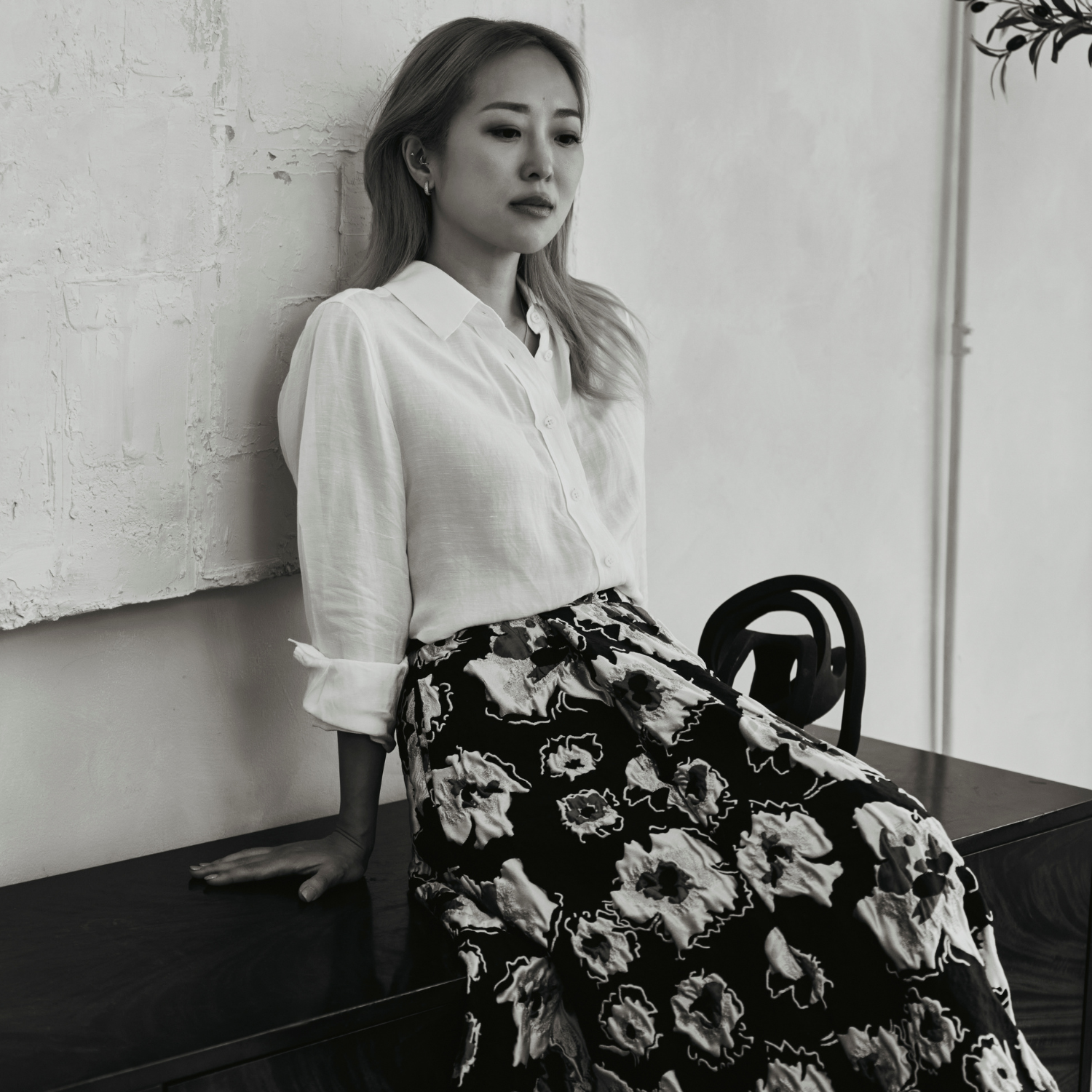 A black and white photo of a woman in a white shirt and a patterned skirt, perched on a sideboard