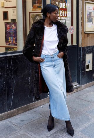 A denim skirt outfit is shown in a photo of a woman on the sidewalk wearing a black fur coat over a white t-shirt with a denim maxi skirt, black tights, and black pointed pumps