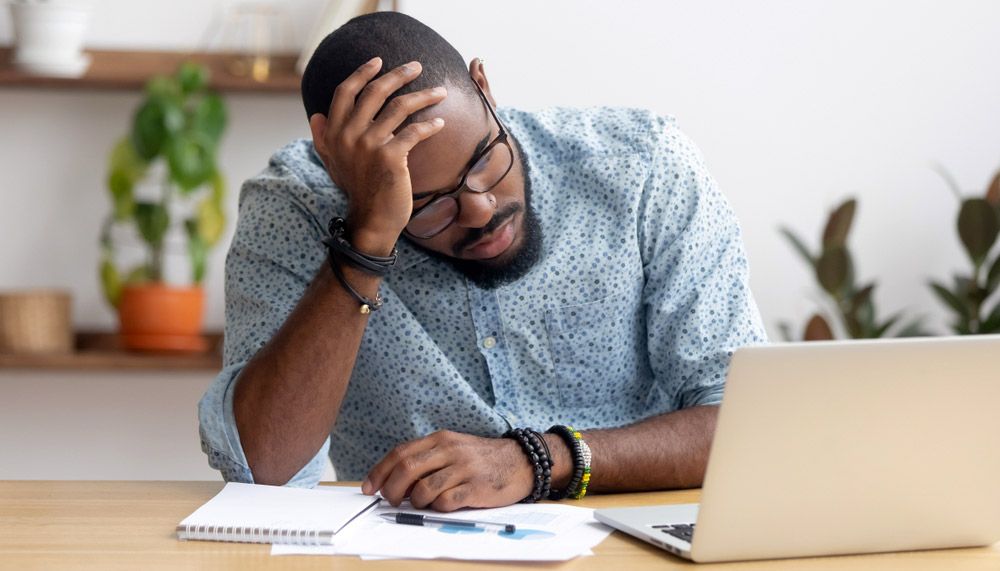 Man with glasses and beard rest head in hand as he stares wearily at laptop screen.