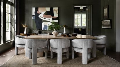 green dining room with large windows and lots of natural light, gray tub chairs, timber table with vase filled with foliage and a large artwork on the wall