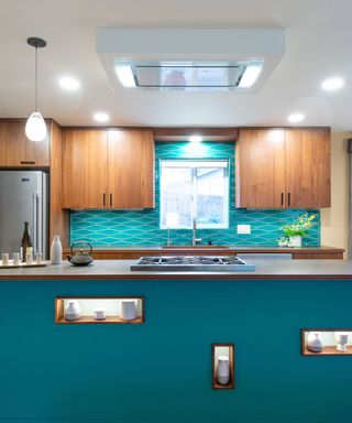 A kitchen with a blue island and splashback, a silver fridge, wooden brown cabinets, and pendant and recessed lighting on the ceiling