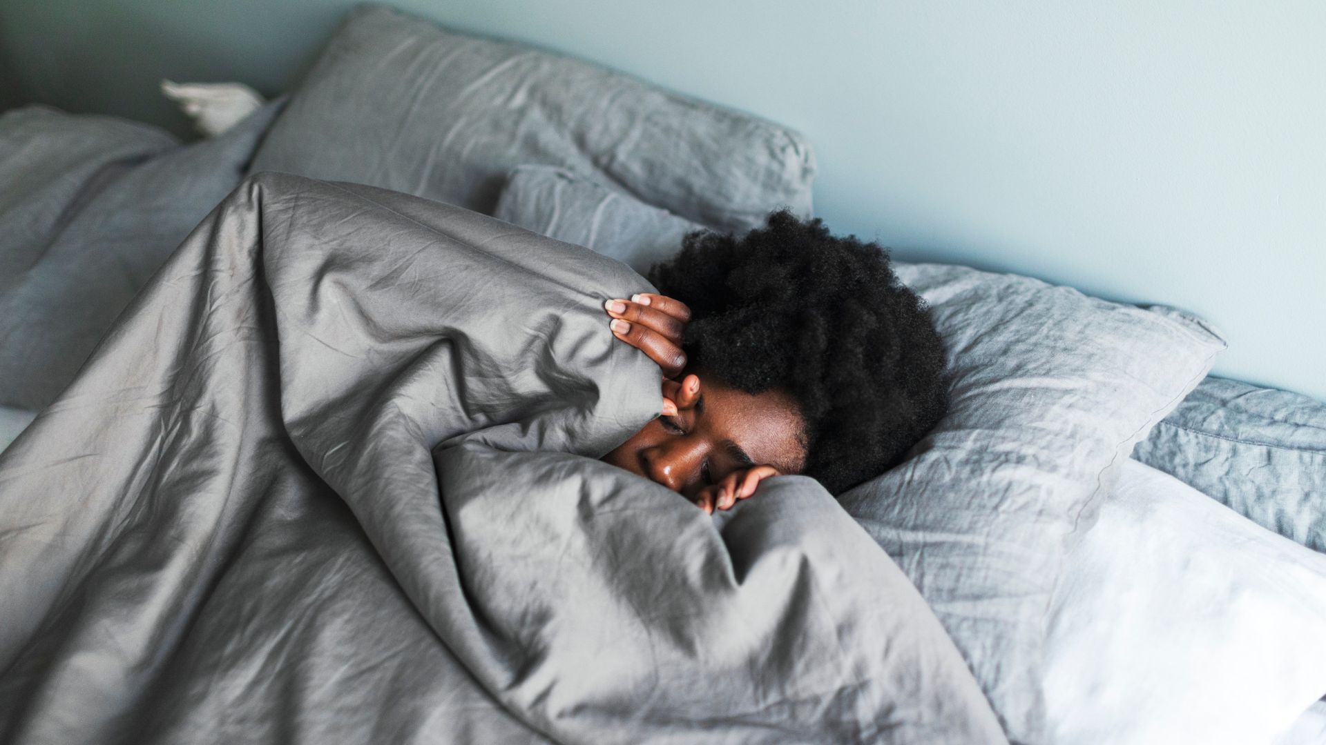 A woman lying in bed with her eyes closed, with a gray duvet pulled up her chin.