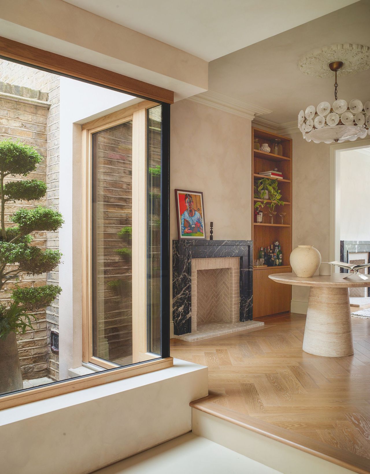 A room with a marble fireplace and large round pedestal table has a corner window with a view to a courtyard and a large potted tree