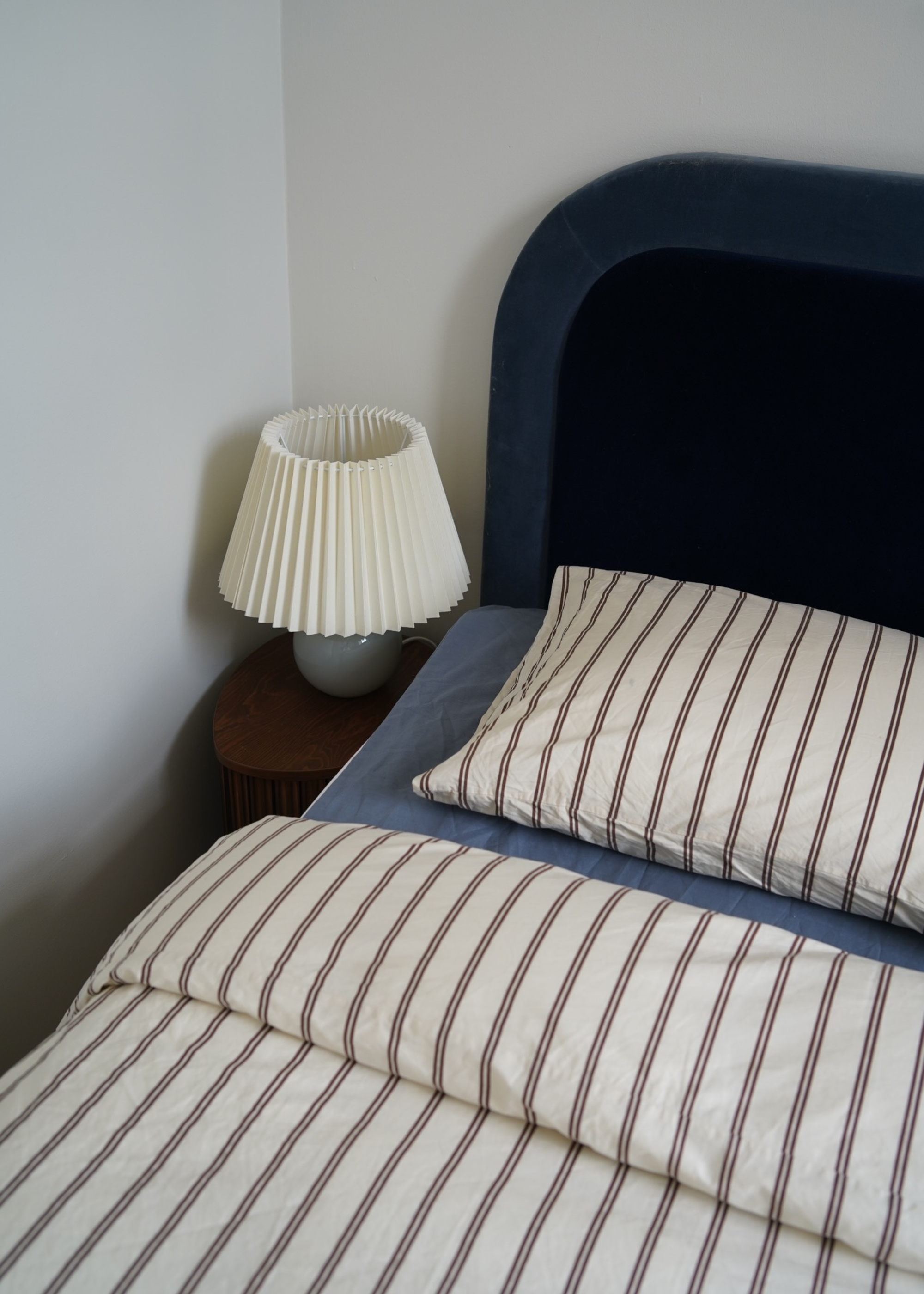 A white bedroom corner with a blue headboard, blue bedding, striped pillow and duvet covers, a small wood side table and a lamp with a pleated shade and a blue base