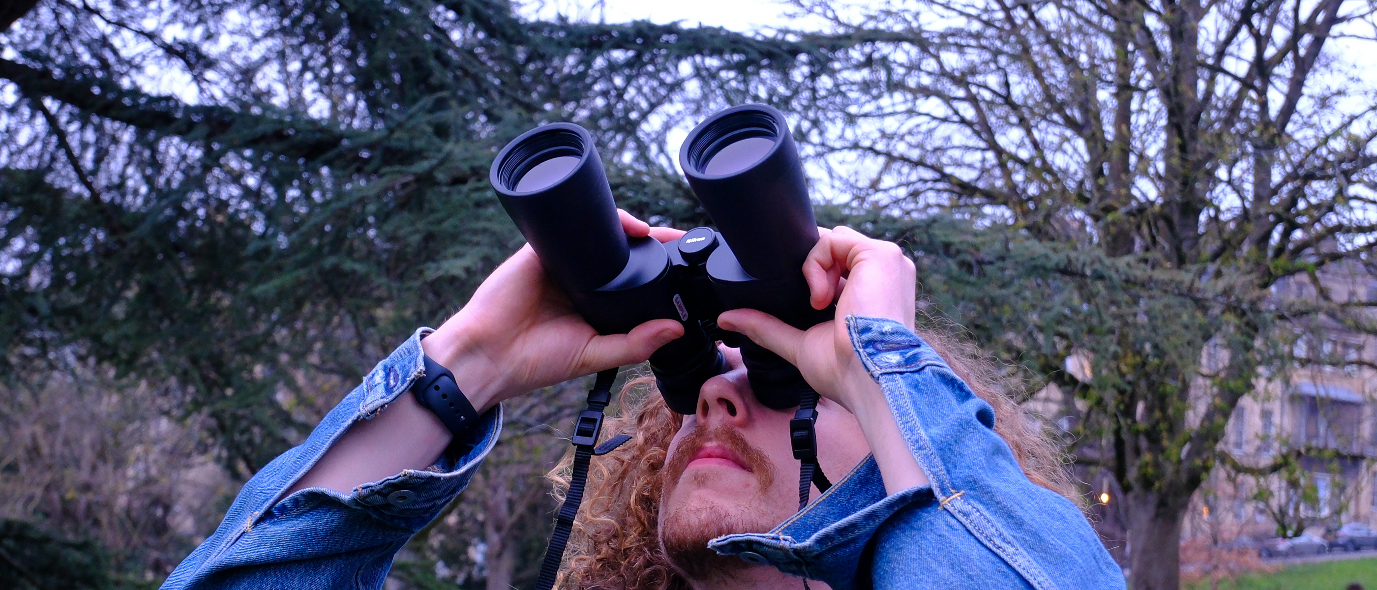 A man looking towards the sky, through the Nikon Action 7x50 binoculars, with trees in the background. 