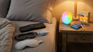 This image shows a warmly lit bedroom with a MUSICOZY sleep headband, Gritin sleep mask and Alpine Silence Sleep earplugs resting on the sheets. On the nightstand next to the bed is a lamp, JALL Sunrise alarm clock, BreatheRight nose strips, and a box with L LOHAS LED amber lightbulbs. The image has been edited by Gemini