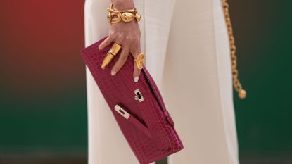 Guest seen outside Elie Saab show wearing beige jumpsuit, gold Schiaparelli belt and bracelet and rings, burgundy crocodile Hermes Kelly clutch with French crème nails
