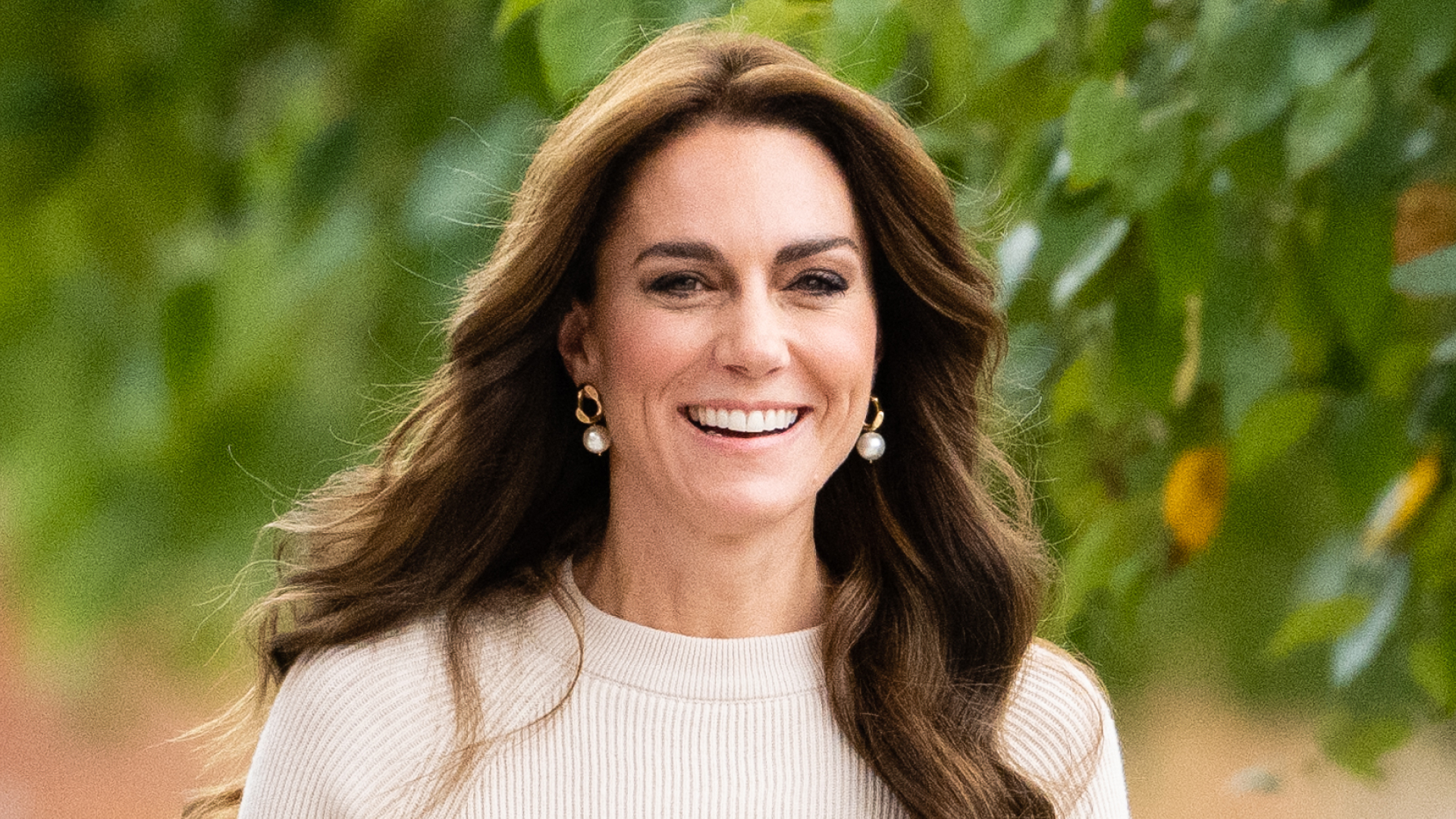 Kate Middleton has long brunette wavy hair and wears a cream sweater and pearl earrings