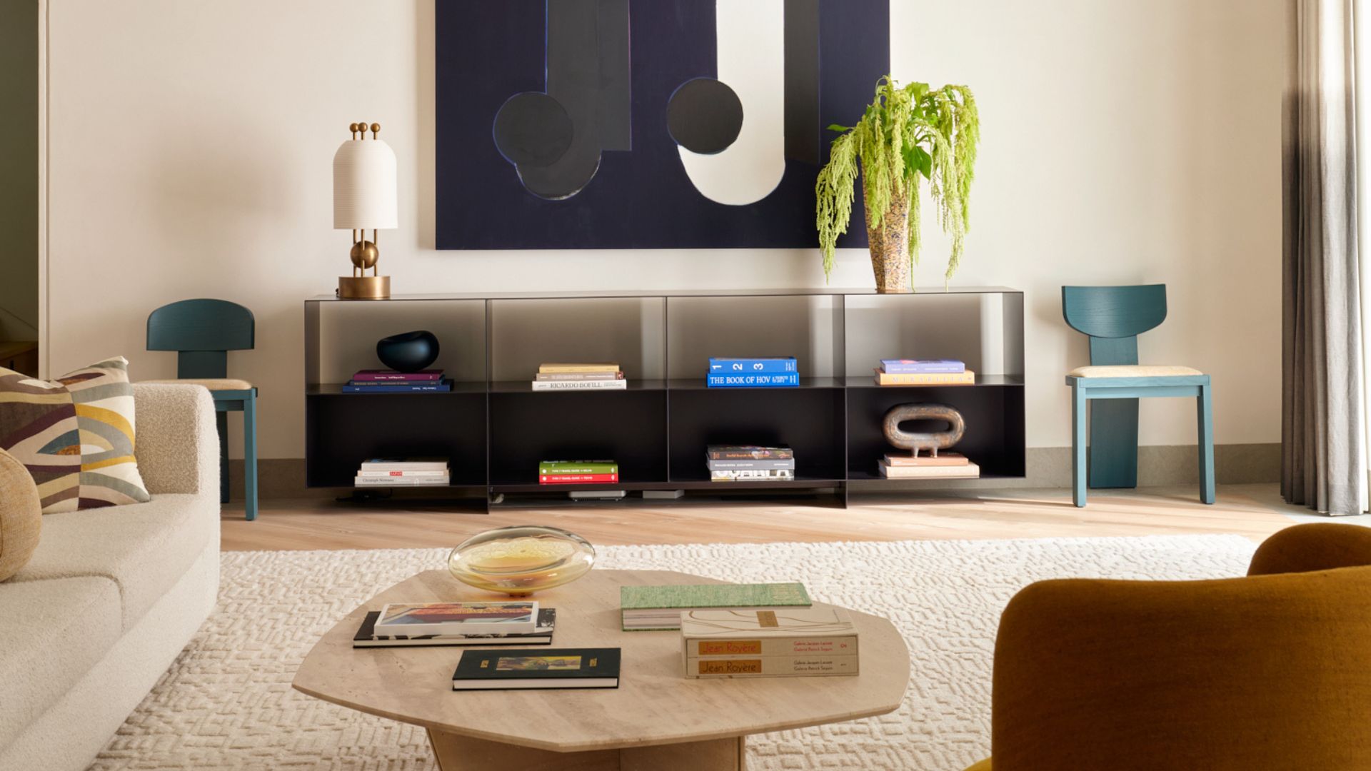 A living room with a black storage case featuring colorful books and ceramics, with a vase of green stems and a gold and white table lamp on top, by a large hanging painting flanked by a pair of teal chairs. Also in the room is a white couch with patterned cushions, a wood coffee table, and a brown armchair on a cream area rug