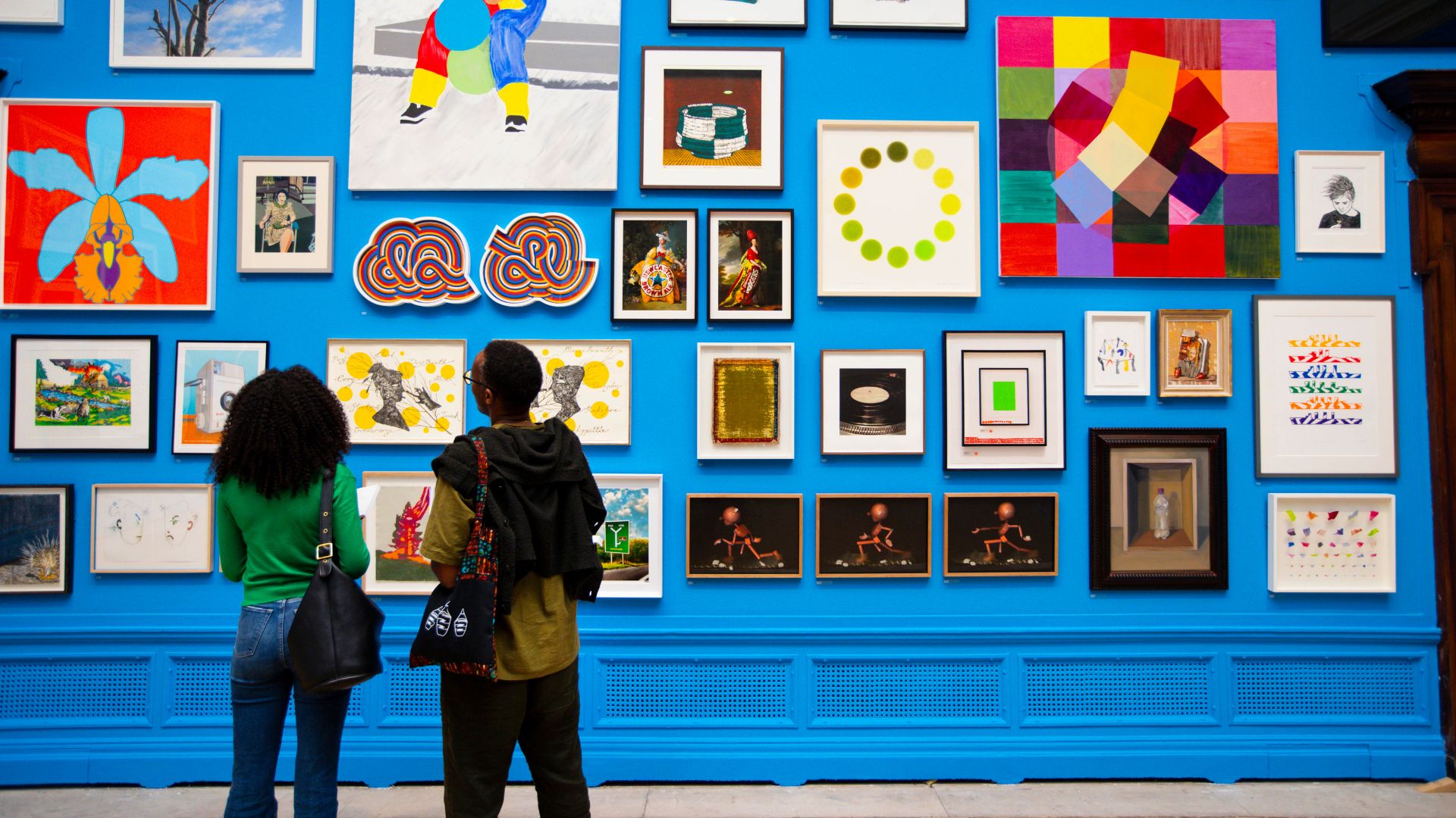an image of two people looking up at a wall of art. The wall is painted bright blue an there are around twenty pieces of art hanging on it, all different sizes and patterns