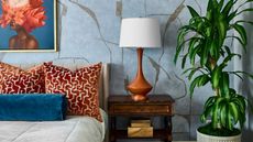 bedroom with blue wallpaper, artwork over bed, red patterned pillows, a red lamp on a timber nightstand, and a potted plant