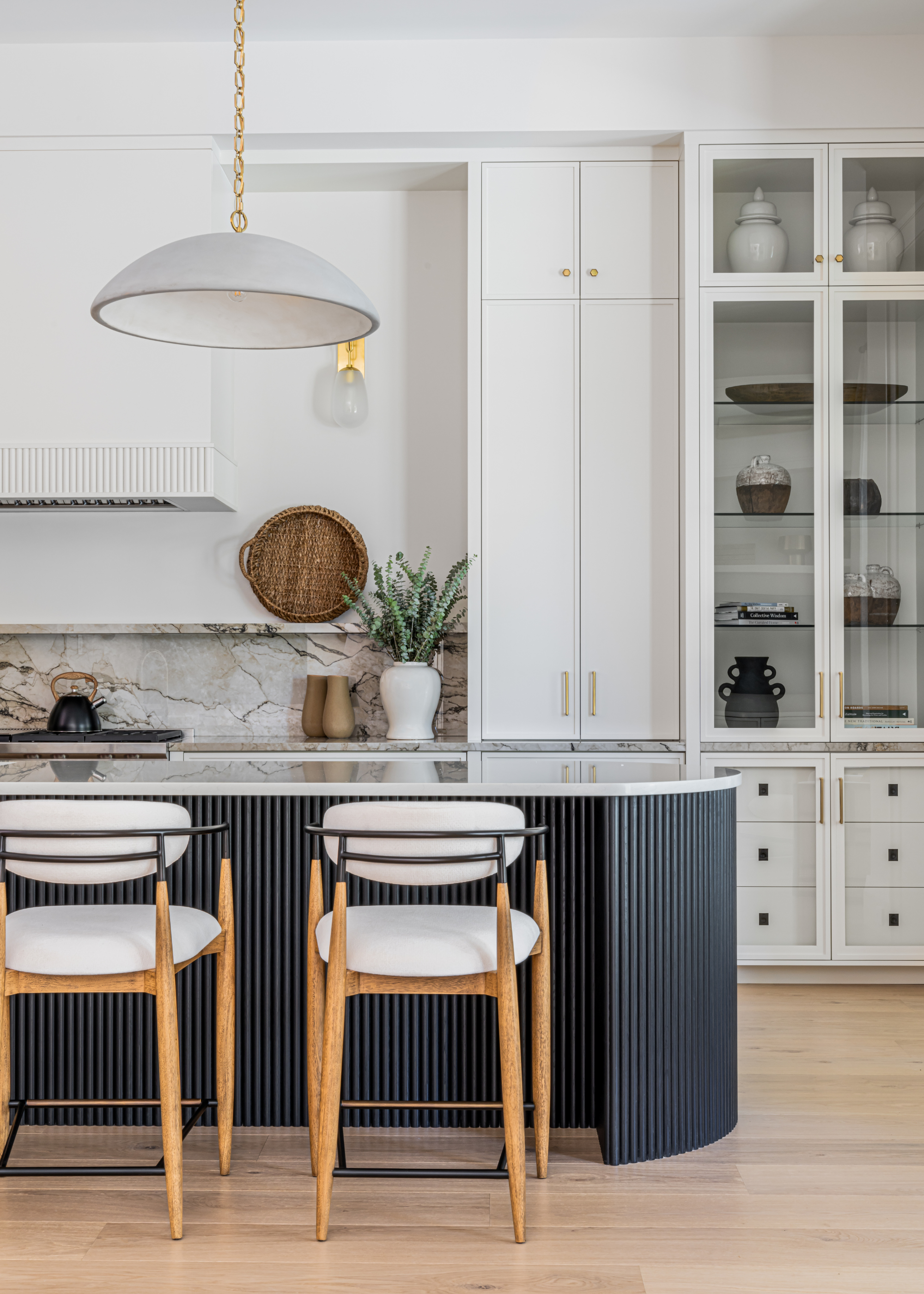 A white kitchen with a marble backsplash, a white cabinet with glass windows, a hanging pendant lamp, a ridged kitchen island with a marble top and a pair of wood stools