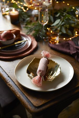 Christmas table gift ideas, elegant table setting with pink handmade cracker, gold and rust color scheme, garland in background with fairy lights