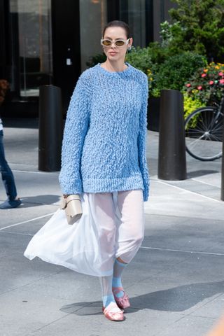 A woman wears a blue textured sweater, a sheer skirt, ballet flats, and beige sunglasses.