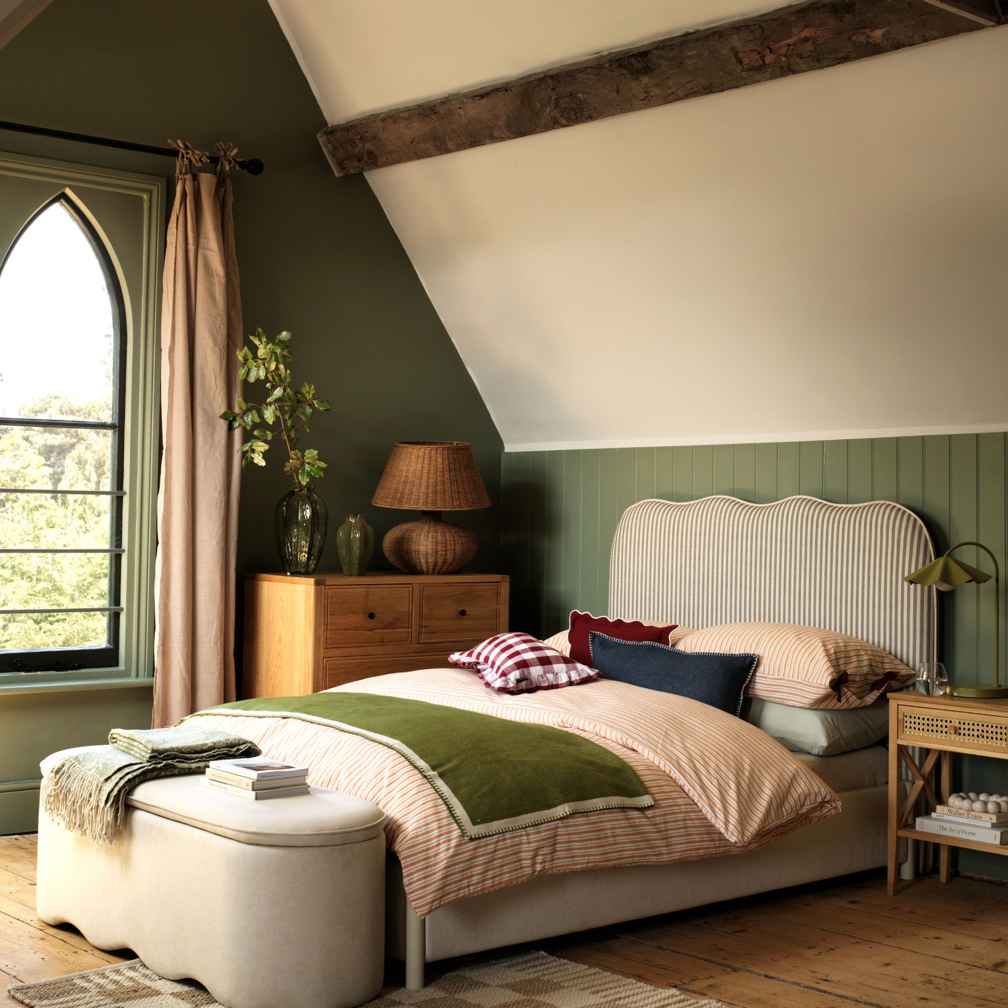 Green bedroom with wooden chest, rattan large lamp and bedside table. Striped headboard with red patterned bedding and green blanket. Natural ottoman at bottom of bed