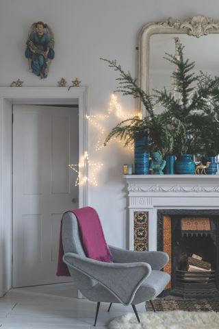Christmas living room with fir tree on the mantel, mirror, blue vases, ornaments, two LED stars, open fire, grey contemporary armchair with pink blanket