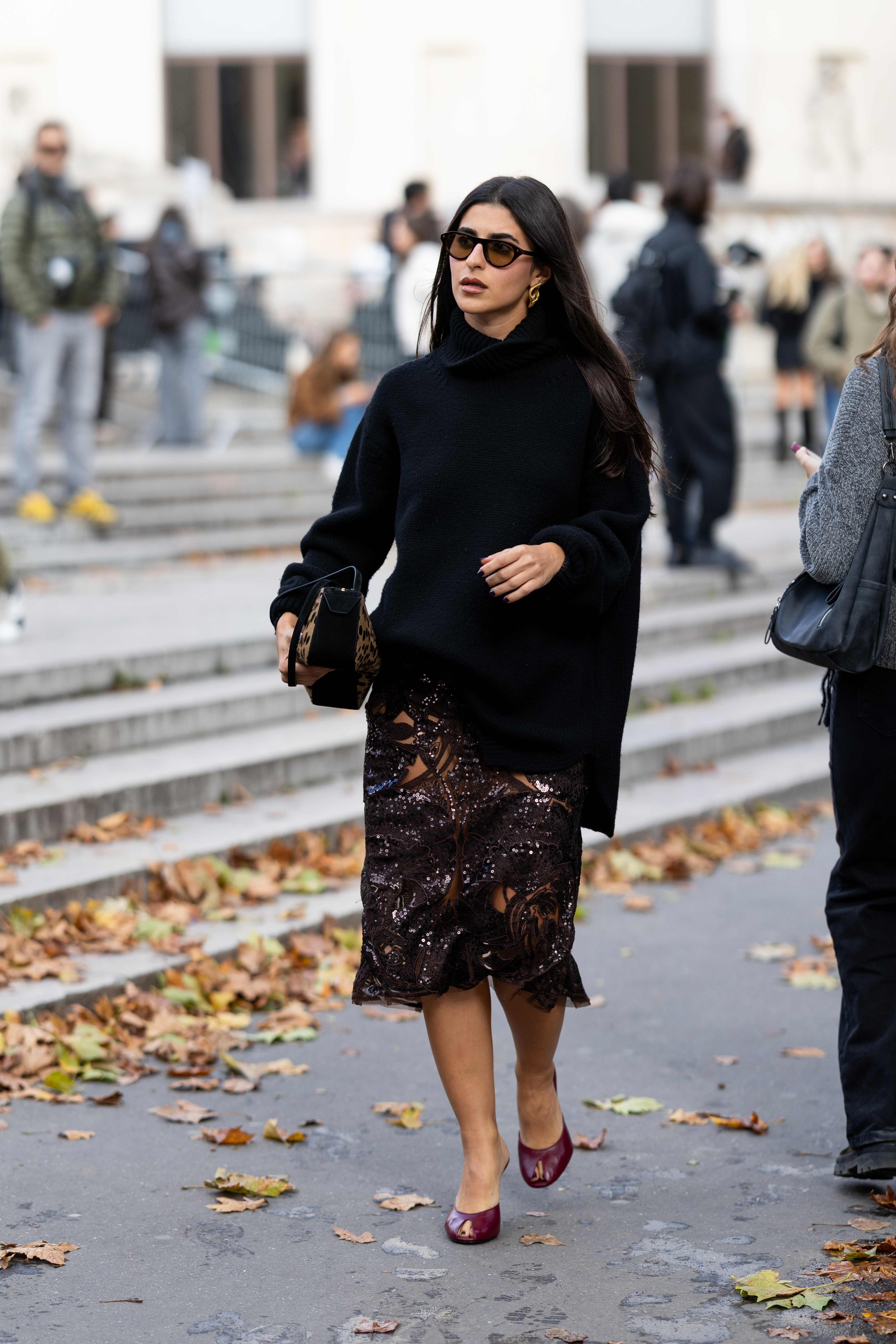 A woman wears a black sweater, brown sequin skirt, burgundy heels, and a leopard bag.