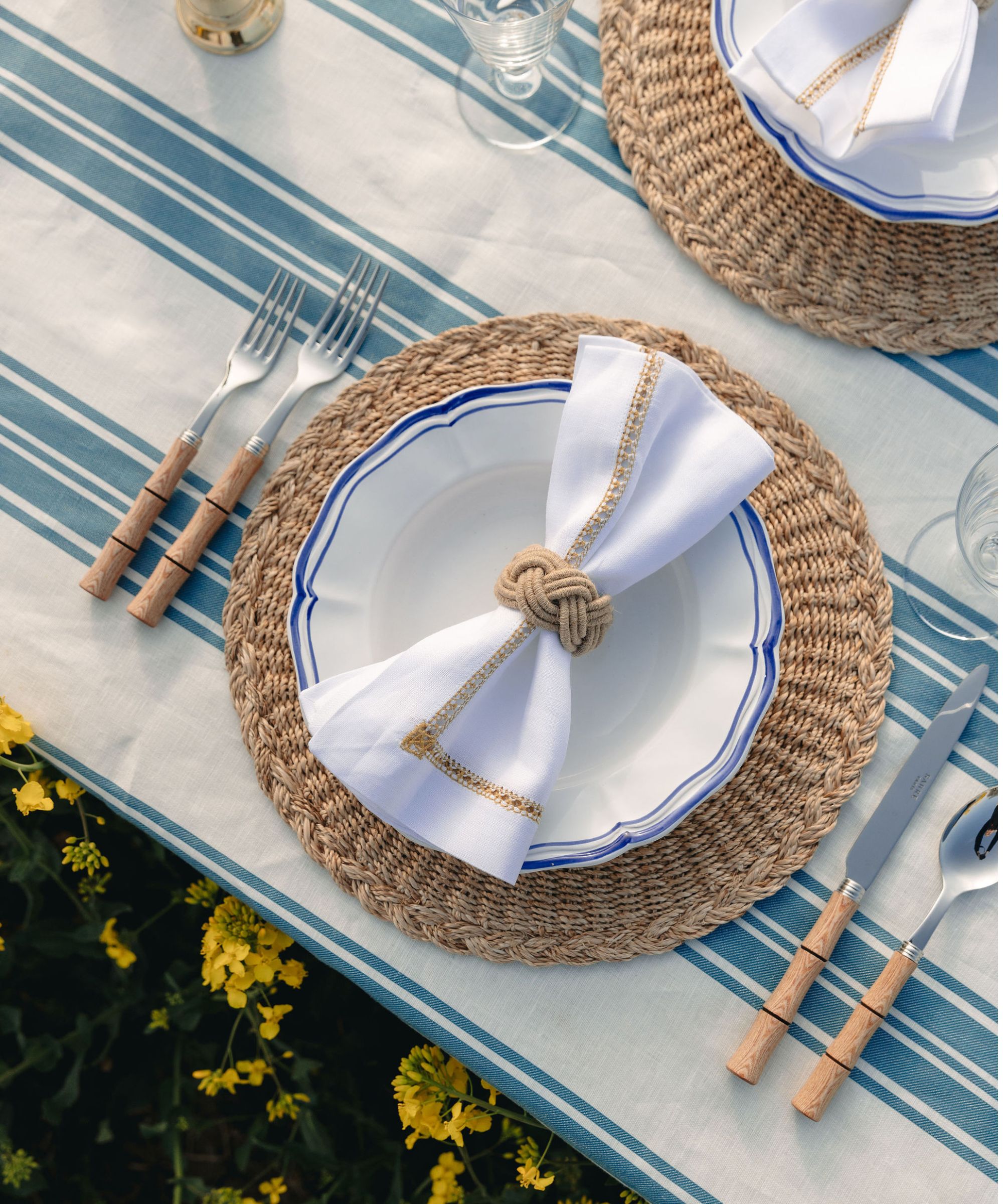 long table with a blue and white striped tablecloth, rattan round charger, bamboo flatware and white napkin