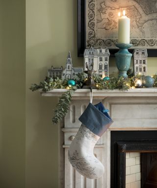 Christmas fireplace decor with foliage, a pop up festive town display, a white stocking and fairy lights