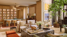 a great room with a large marble coffee table with several books and trinkets on top, two white accent chairs, two burnt orange stools, and a large potted plant. Behind the sitting area is a dining room with a large baby blue coffee table, several dining chairs, and a wall full of abstract framed prints