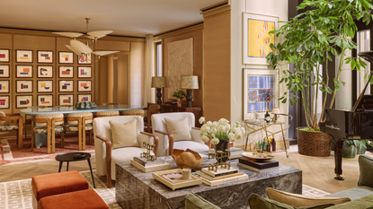 a great room with a large marble coffee table with several books and trinkets on top, two white accent chairs, two burnt orange stools, and a large potted plant. Behind the sitting area is a dining room with a large baby blue coffee table, several dining chairs, and a wall full of abstract framed prints