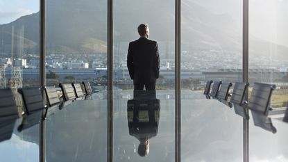 A businessman stands in front of tall windows in an office conference room and looks out.