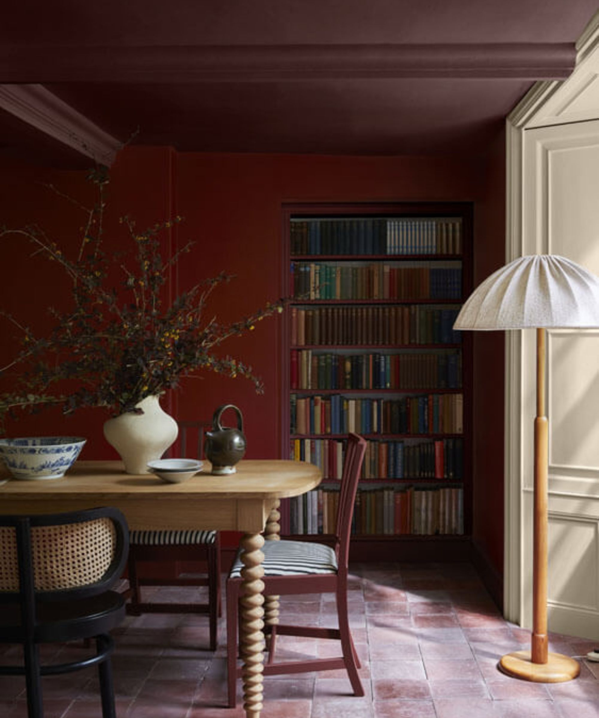 Color drenched red dining room with a wooden dining table and decorated with a vase of fall foliage and a wooden lamp with white shade