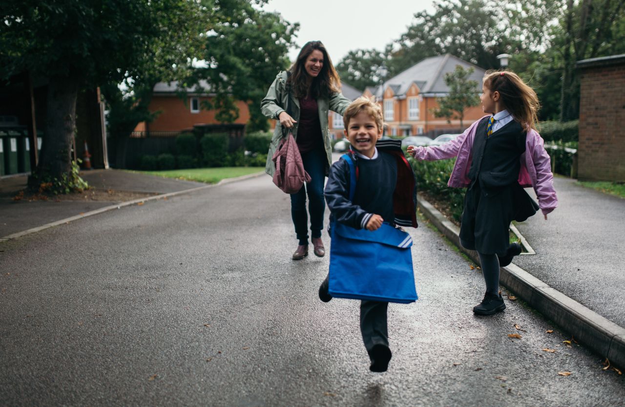 A mum chasing her kids down the road as rush to school in their uniforms, laughing 