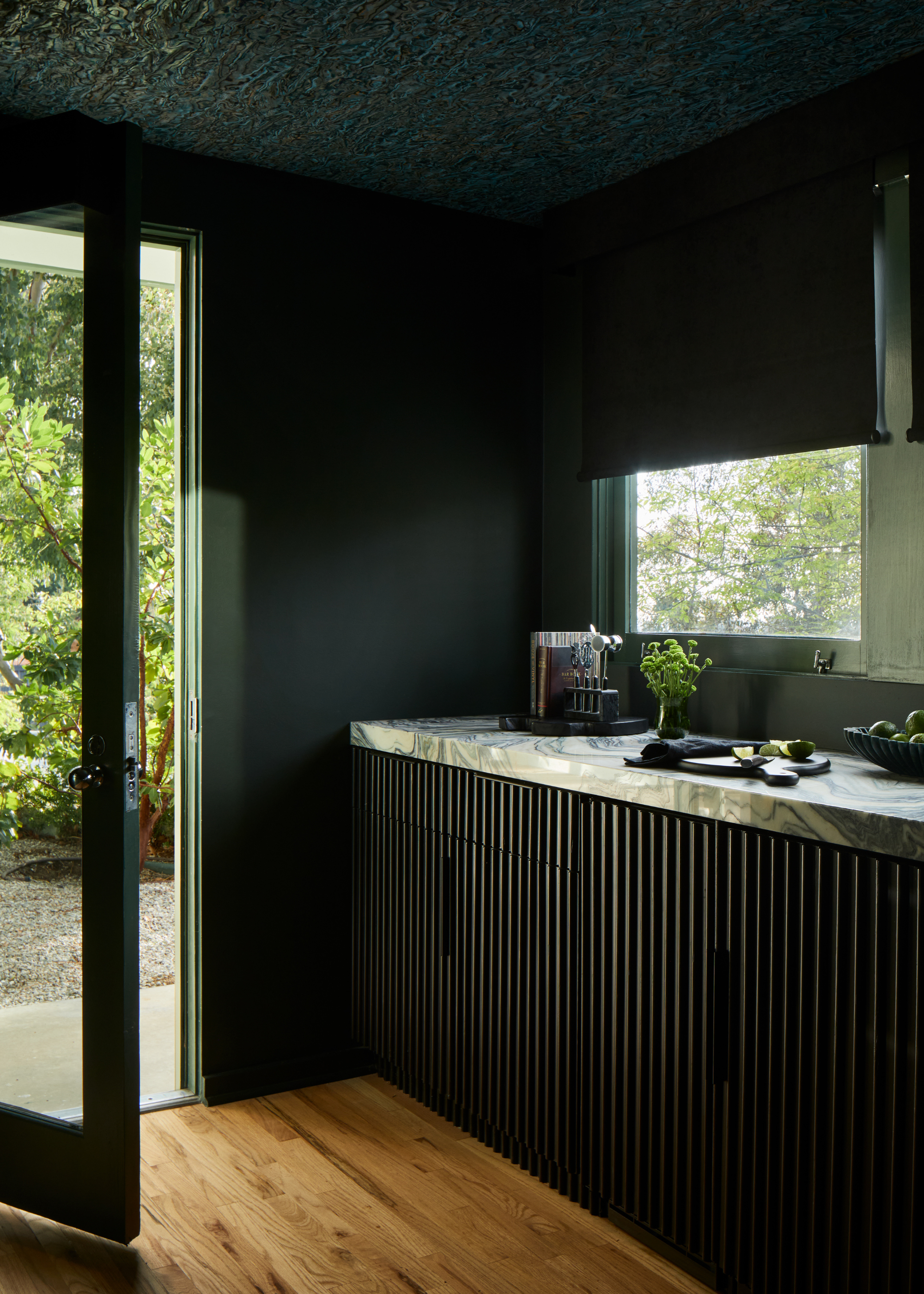 A dark green kitchen with a door leading to a backyard. In the kitchen, there's a stone island, with a chopping board, avocadoes, a knife, a potted herb and a bowl of fruit