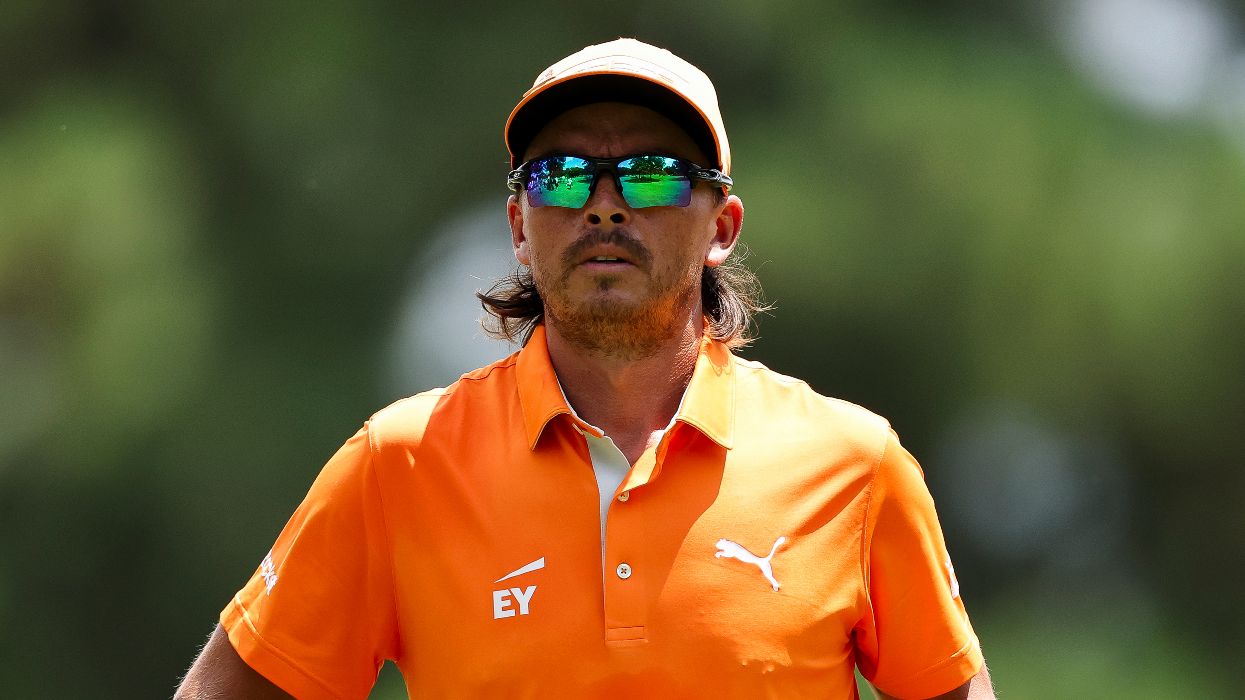 Rickie Fowler wearing an orange polo and cap with green sunglasses