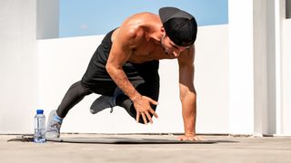 Man outdoors topless in hat and shorts in a high plank with right arm and left leg lifted in the air