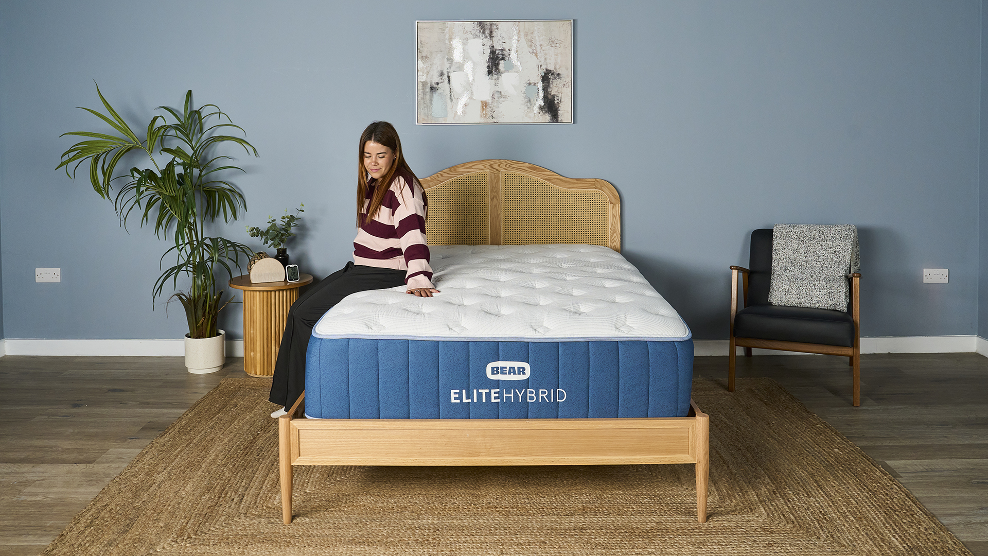 A person is sitting on the side edge of the mattress, looking down toward the floor. The mattress shows minimal visible compression, highlighting the strength of its edge support. The blue side panel of the mattress is clearly visible.