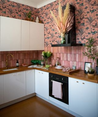 kitchen with white kitchen cabinets and wall units, wooden countertop, black cooker hood, pink backsplash tiles and wallpaper