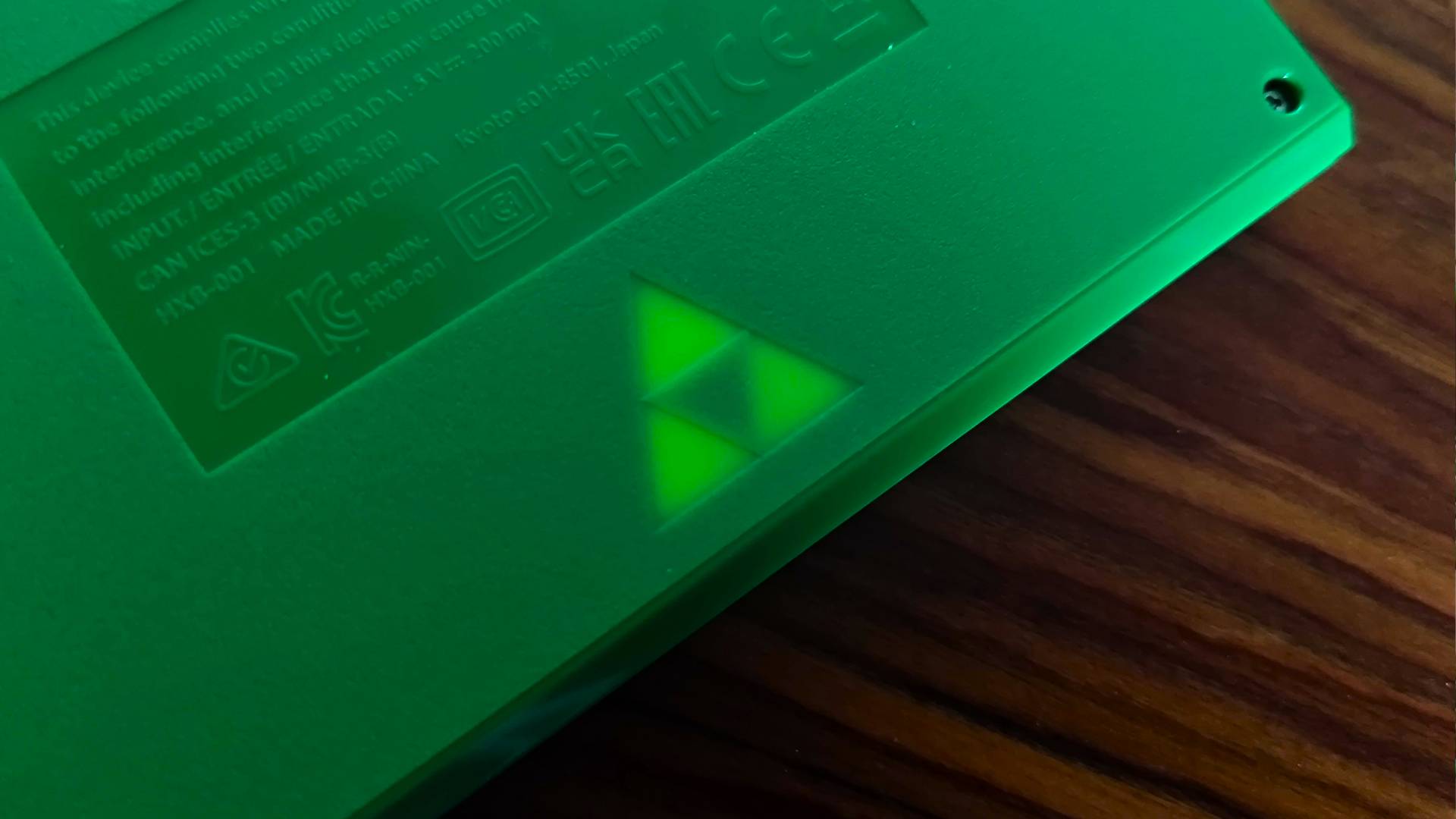 Close up of Zelda Game and Watch glowing Triforce on back of handheld sitting on woodgrain desk.