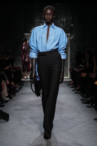 A model wearing a blue button-down shirt tucked into trousers walking in Haider Ackermann's first Tom Ford runway show during Paris Fashion Week.