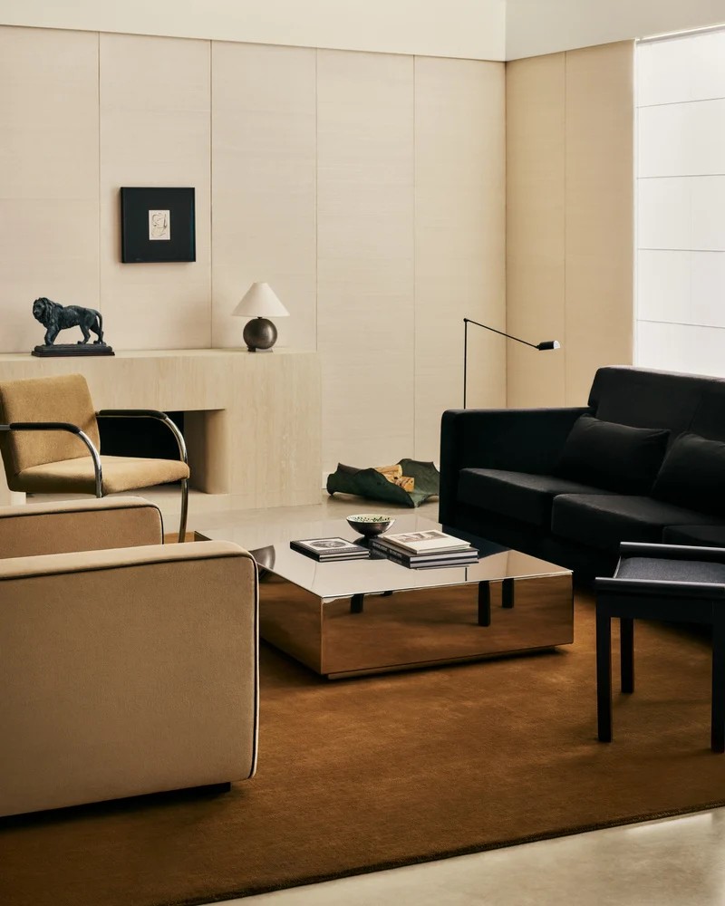 A modern, elegant living room with off-white walls and a caramel brown rug. There are cream, chrome, and black modern furniture pieces around the room.