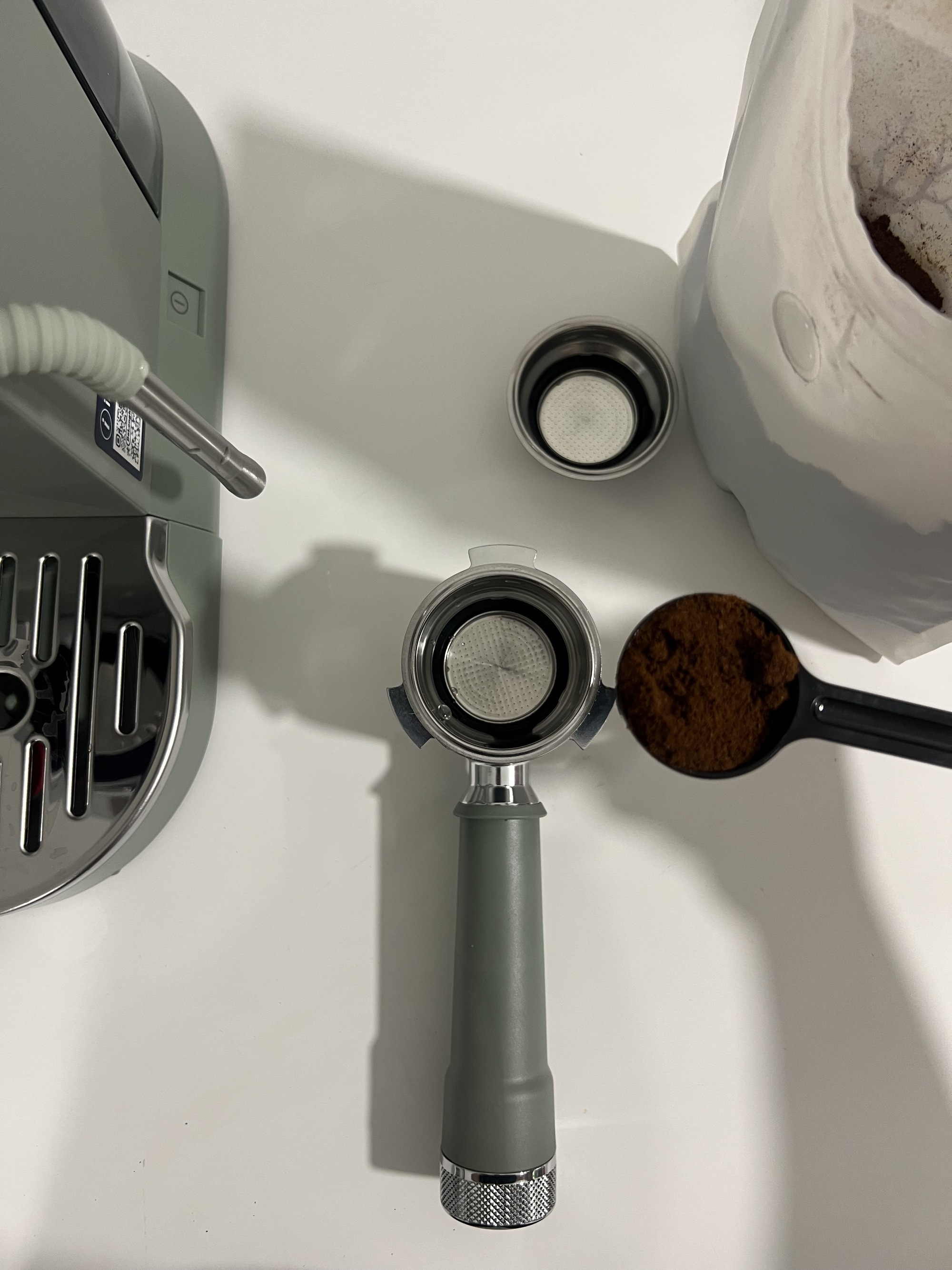 Image of a sage green portafiller on a white countertop. There is an open bag of espresso grounds and a full measuring scoop hovering over the portafiller.