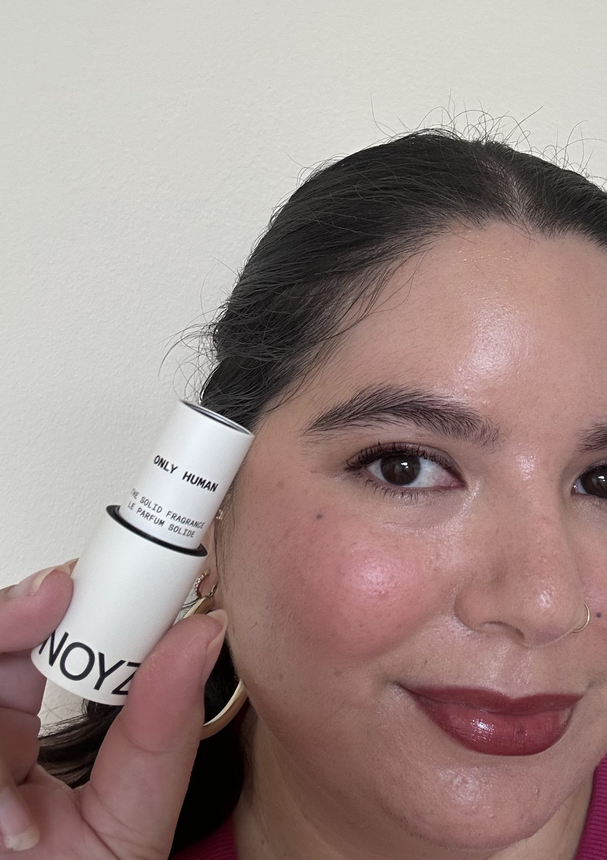 Holding the white NOYZ Only Human solid fragrance stick next to my face, close-up of glowy skin and glossy berry lips
