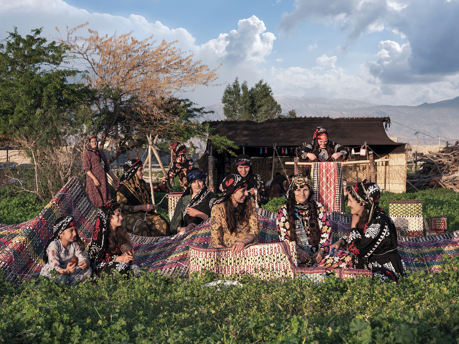 A group of women dressed in colorful traditional attire sits on vibrant woven textiles in a lush green setting, with hills in the background