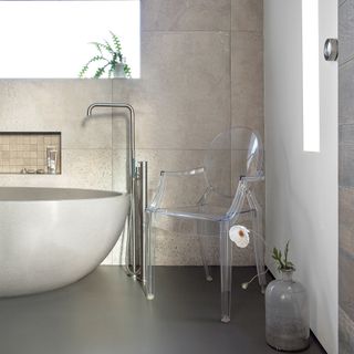 A modern bathroom with large grey wall tiles, bath and transparent chair decor