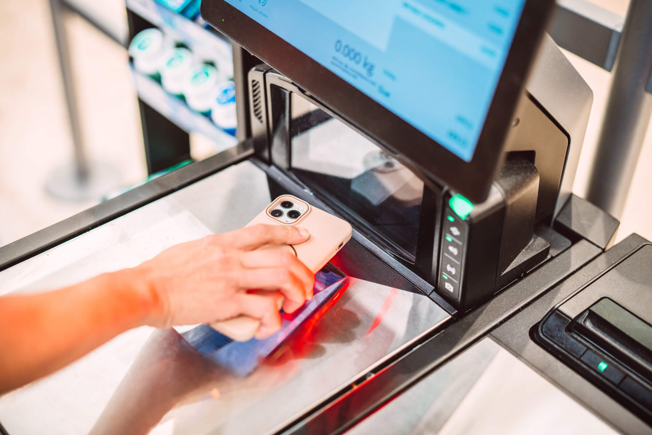 Close-up of a shopper using a smartphone to scan a loyalty card before payment at a supermarket self-checkout.