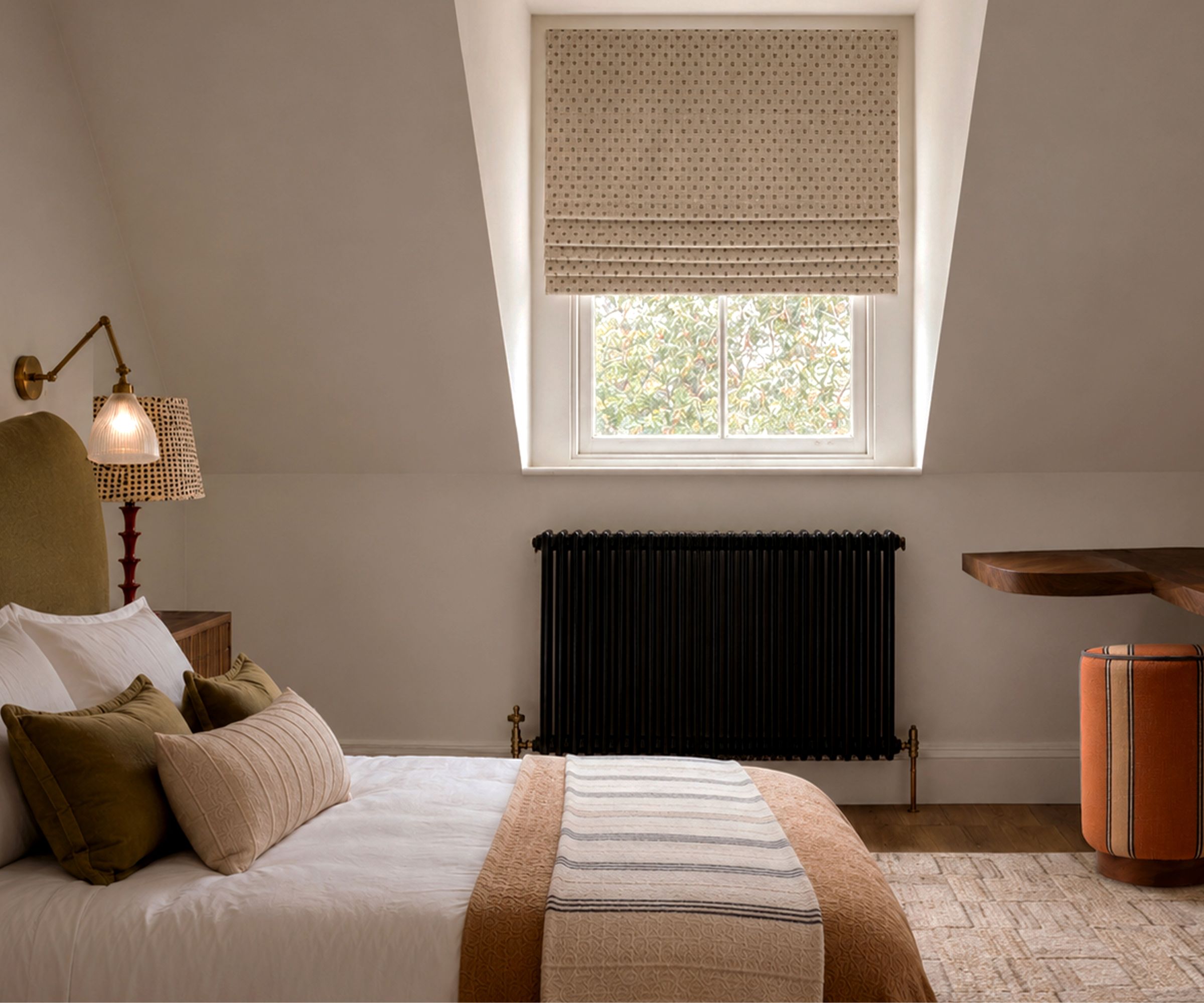 Autumnal bedroom with white walls, a large window with a Roman blind, an olive green bed with neutral bedding and a wool brown throw, a cream rug, with a wall sconce above, and an orange pouffe.