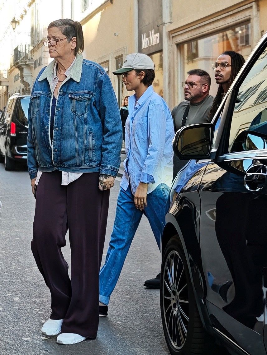 Zendaya walks down the street wearing a blue button down shirt with black leather loafers and blue jeans.
