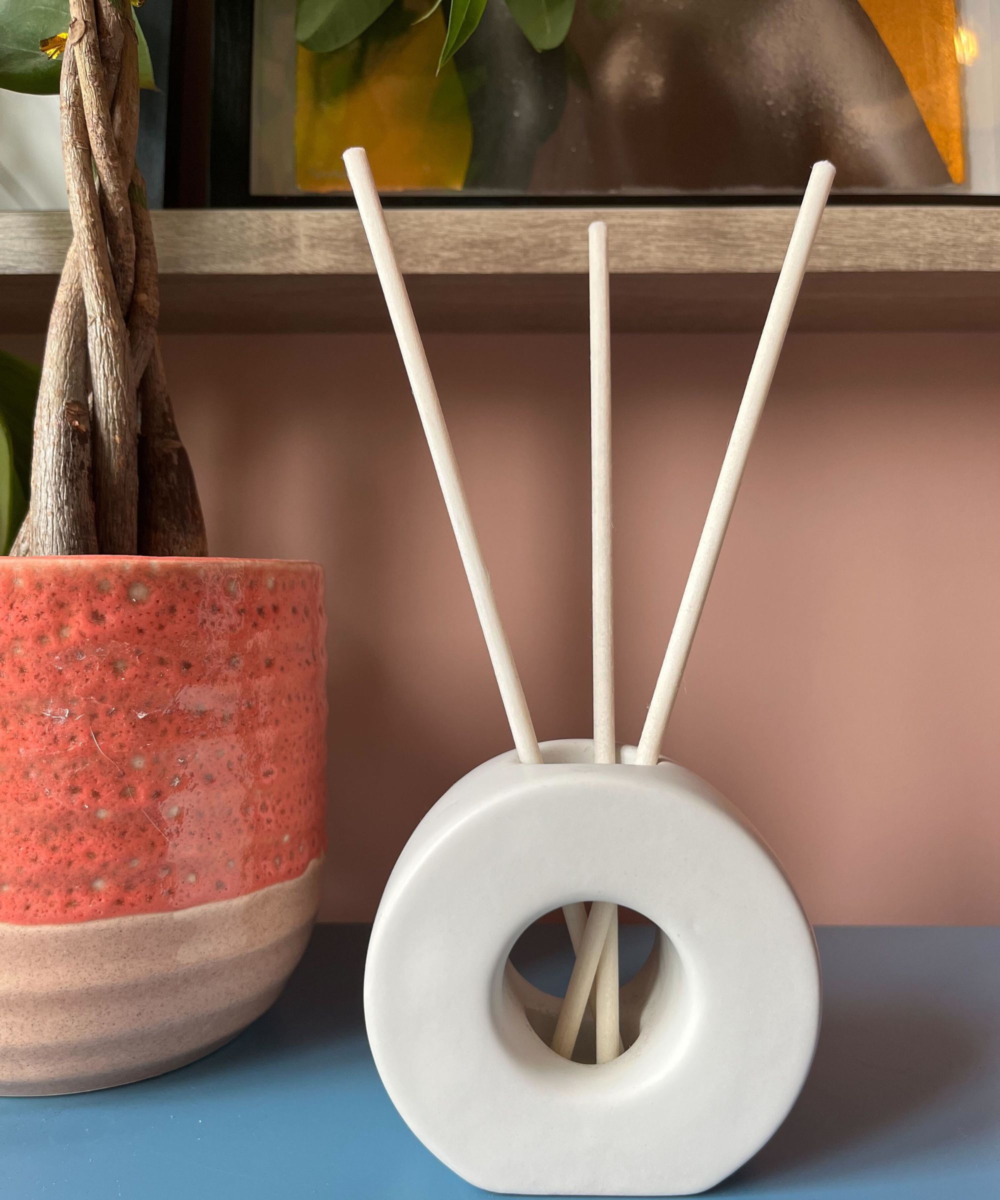 A ScentAir reed diffuser on a blue surface beside a pink plant pot with a small tree growing. Above is a wooden shelf with the bottom half of an artwork shown, and the wall behind is a pale, peachy pink.