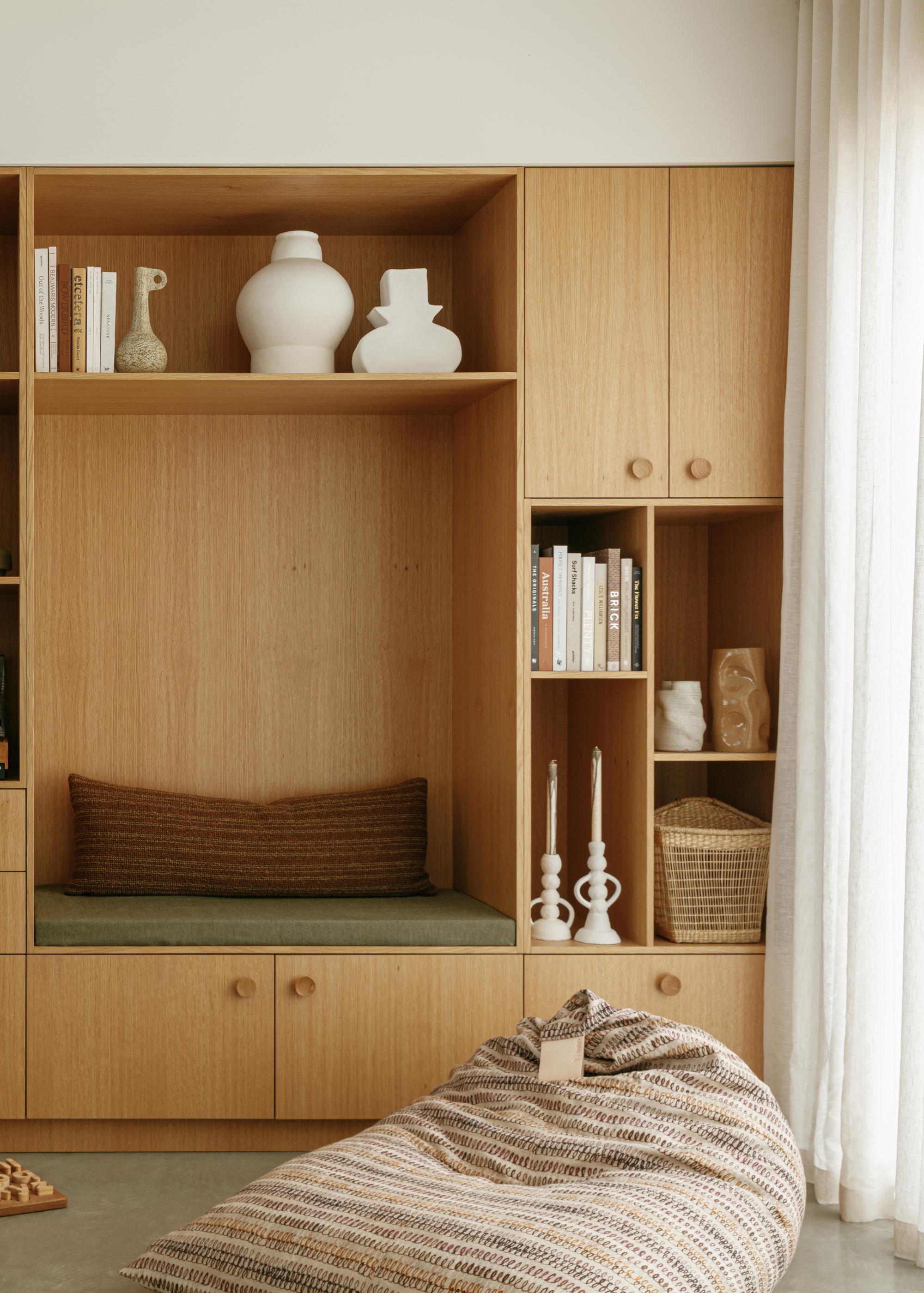 A sitting room with an in-built nook, a wooden cupboard, shelves with candles, vases, and books, alongside a textured bean bag