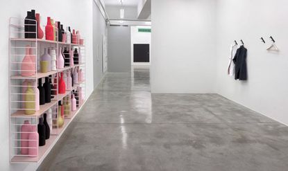 View of Matthew Brannon's show at the Casey Kaplan gallery featuring a pink, wall-mounted shelving unit with bottles in different colours, grey floors and light coloured walls with individual coat hooks, two coats and a hanger on one side