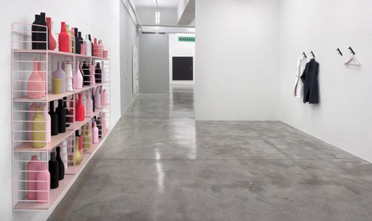 View of Matthew Brannon&#039;s show at the Casey Kaplan gallery featuring a pink, wall-mounted shelving unit with bottles in different colours, grey floors and light coloured walls with individual coat hooks, two coats and a hanger on one side