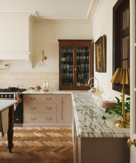 The corner of a Victorian kitchen with pink cabinets, cream walls, and an antique wooden cabinet sat on the marble countertops, storing glassware and mugs